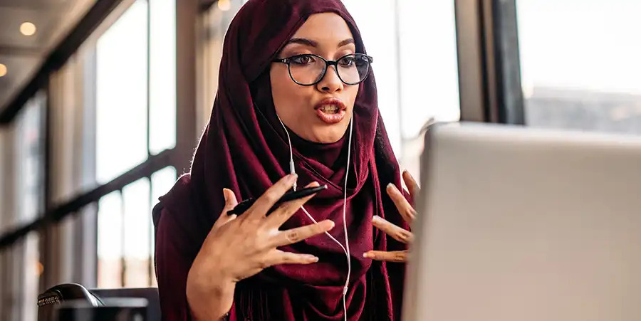 Una mujer con gafas de lectura y un hiyab morado animada mientras habla mirando la pantalla de un ordenador portátil, en una reunión en línea.